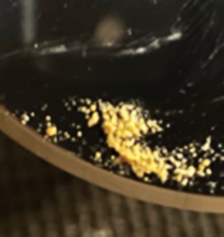 The image shows a close-up of small, yellowish particles scattered on a dark, reflective surface, possibly a black dish or pan. The particles vary in size, with some clustering together, creating a gritty texture that stands out against the smooth, shiny background.